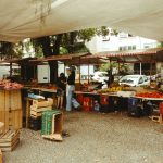 Feira de alimentos ao ar livre, com barracas de produtos frescos sob uma cobertura e circulação de visitantes.