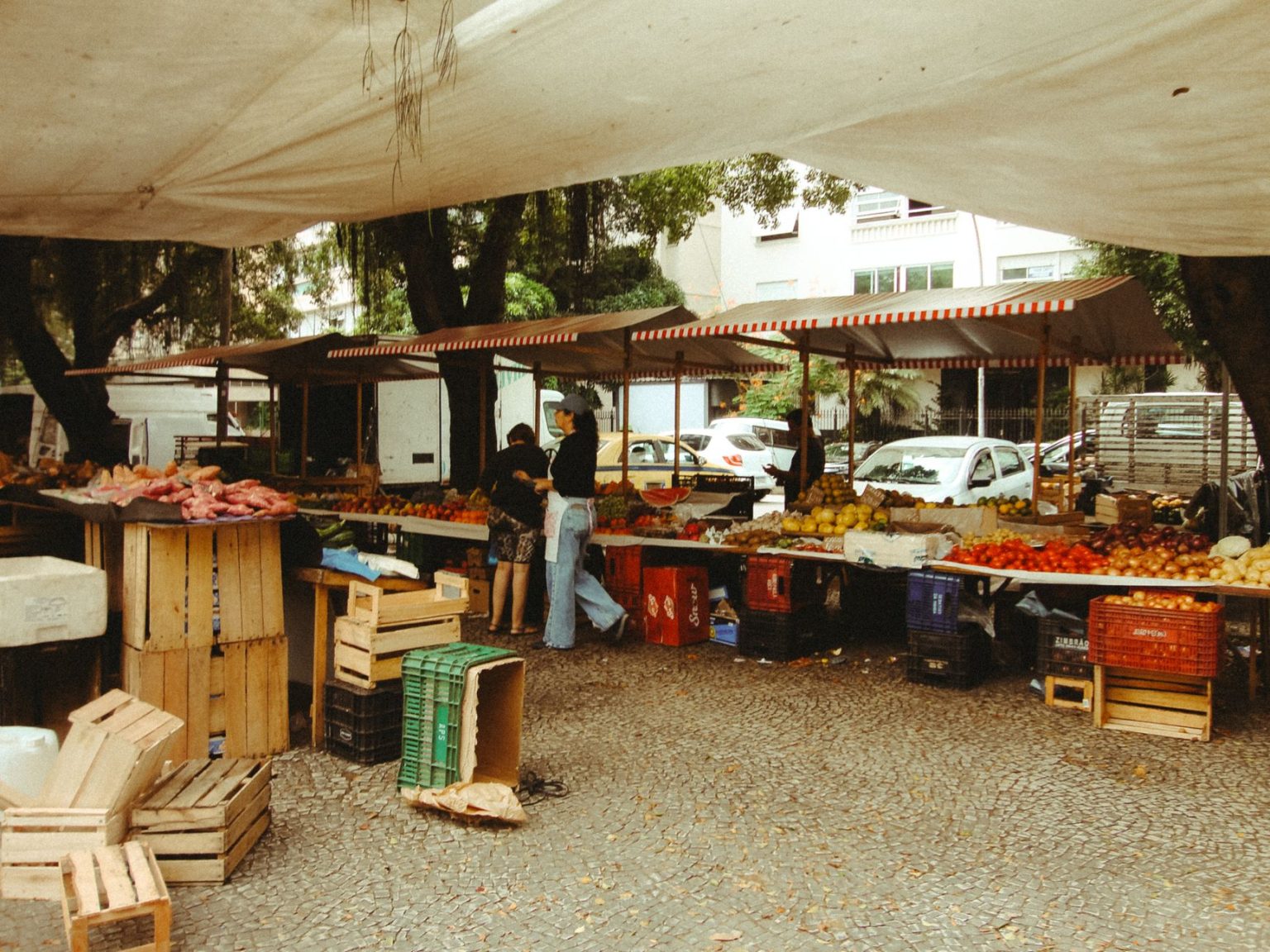 Feira de alimentos ao ar livre, com barracas de produtos frescos sob uma cobertura e circulação de visitantes.