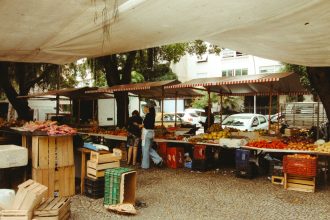 Feira de alimentos ao ar livre, com barracas de produtos frescos sob uma cobertura e circulação de visitantes.