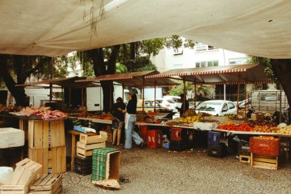 Feira de alimentos ao ar livre, com barracas de produtos frescos sob uma cobertura e circulação de visitantes.