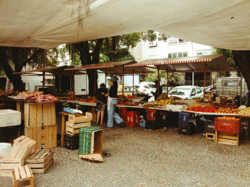 Feira de alimentos ao ar livre, com barracas de produtos frescos sob uma cobertura e circulação de visitantes.