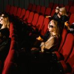 Pessoas usando óculos 3D assistem a um filme e comem pipoca em uma sala de cinema com poltronas vermelhas.
