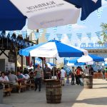 Pessoas aproveitam um festival ao ar livre, com mesas, comidas, bebidas, guarda-sóis coloridos e bandeirolas.