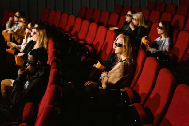 Pessoas usando óculos 3D assistem a um filme e comem pipoca em uma sala de cinema com poltronas vermelhas.