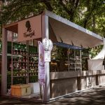 Barraca de vinhos em um evento ao ar livre, cercada por árvores e visitantes.