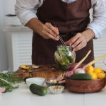 Pessoa usando uma pinça para preparar vegetais em conserva dentro de um pote de vidro, sobre uma mesa de cozinha.