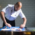 Chef de casaco branco filetando um peixe sobre uma tábua azul em uma cozinha profissional.