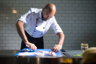 Chef de casaco branco filetando um peixe sobre uma tábua azul em uma cozinha profissional.