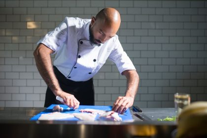 Chef de casaco branco filetando um peixe sobre uma tábua azul em uma cozinha profissional.