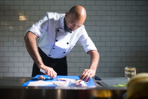 Chef de casaco branco filetando um peixe sobre uma tábua azul em uma cozinha profissional.