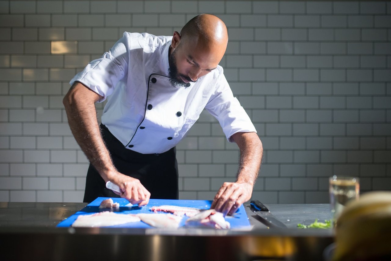 Chef de casaco branco filetando um peixe sobre uma tábua azul em uma cozinha profissional.