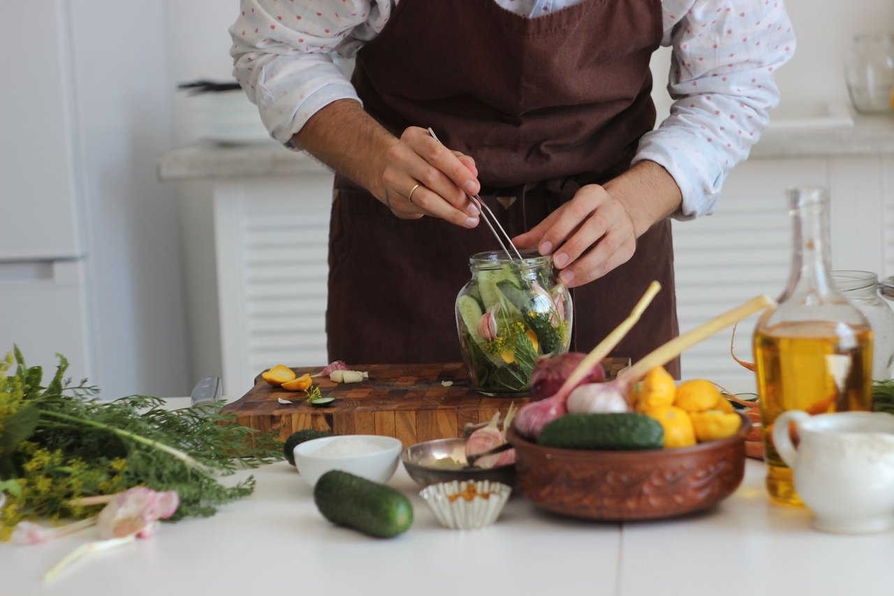 Pessoa usando uma pinça para preparar vegetais em conserva dentro de um pote de vidro, sobre uma mesa de cozinha.