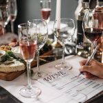 Mesa de degustação com taças de vinho, queijos e anotações de avaliação em um ambiente interno.